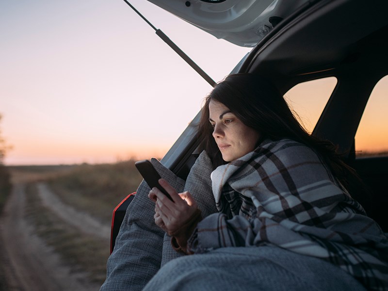 Kvinna sitter i bagaget på bilen med öppen lucka med en filt om axlarna mot den kyliga kvällsluften och tittar på sin telefon