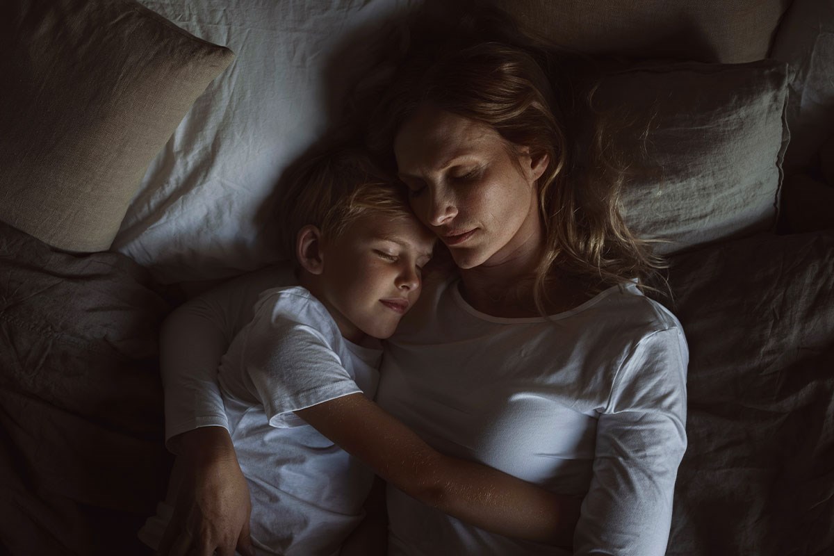 Mor och son som sover tillsammans i en säng, inlindade i vita lakan. Båda ser avslappnade och fridfulla ut